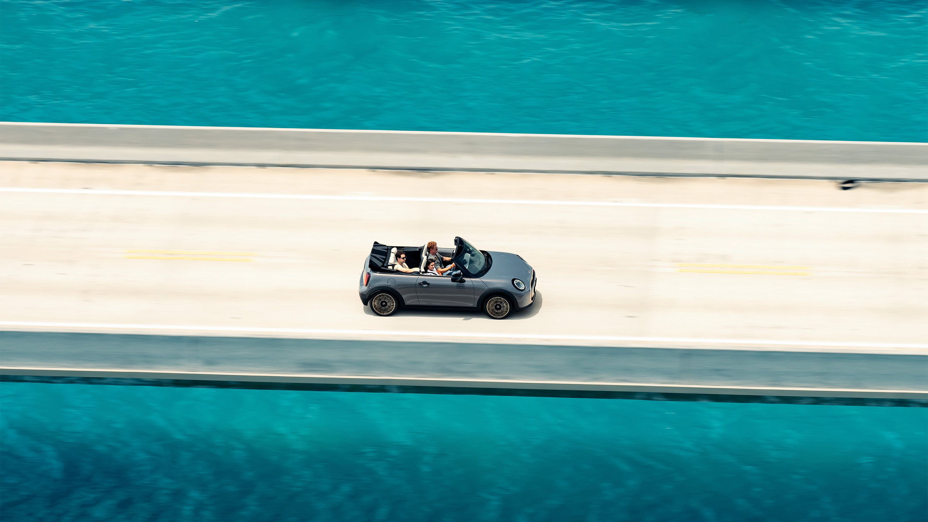 Overhead shot of the new MINI Cooper Convertible with an open top and three occupants, being driven down a road that has turquoise blue water at either side.